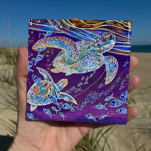 May include: Square artwork featuring two sea turtles swimming in a purple ocean, surrounded by small fish. The turtles have intricate patterns in blue, green, and orange. The background suggests a sunny day at the beach.