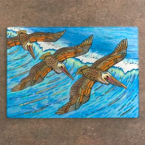 May include: A rectangular artwork depicts three pelicans in flight over a blue ocean wave. The pelicans have brown and orange wings and long beaks. The artwork is painted with blue, white, and brown colors.