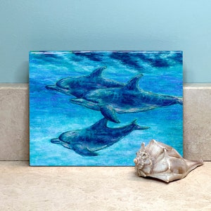 May include: A painting of three dolphins swimming in a blue ocean. The dolphins are painted in shades of blue and gray, and the water is a light blue color. The painting is done in a realistic style.