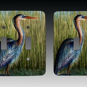 May include: A pair of light switch covers featuring a watercolor painting of a blue heron standing in tall green reeds.