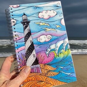May include: A spiral bound notebook with a colorful illustration of a lighthouse, waves, and birds against a blue sky. The cover features a black and white striped lighthouse with a white door and windows.