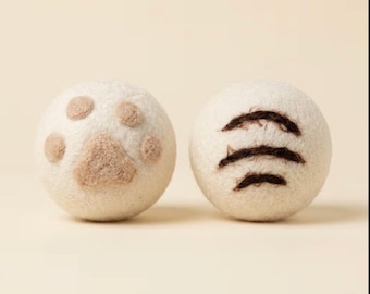 May include: Two round, off-white felted wool balls. The ball on the left features a light tan needle-felted paw print design. The ball on the right displays three horizontal, dark brown needle-felted stripes across its surface.