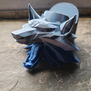 Może przedstawiać: 3D-drukowana rzeźba głowy wilka w odcieniach szarości i niebieskiego. Głowa wilka jest szczegółowa z geometrycznymi fasetami i ostrymi kątami, z niebieską sekcją na dole. Rzeźba stoi na teksturowanej powierzchni.