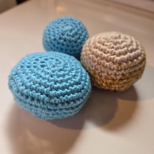 May include: Three spherical objects crafted with crochet yarn. Two of the spheres are a bright, solid light blue color, while the third sphere features a gradient pattern transitioning from a cream white top to a golden yellow base.