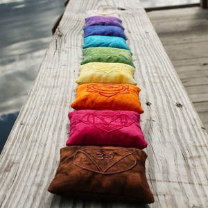 Könnte beinhalten: Eine Reihe farbenfroher, rechteckiger Kissen mit gestickten Designs, angeordnet auf einer verwitterten Holzoberfläche. Die Kissen sind in den Farbtönen Lila, Blau, Türkis, Grün, Gelb, Orange, Rosa und Braun gehalten. Der Hintergrund zeigt Wasser und Himmel.