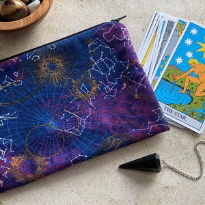 May include: A blue zippered pouch with a constellation print. The pouch is lying next to a deck of tarot cards, with the "The Star" card face up. A black pendulum is also visible in the image.