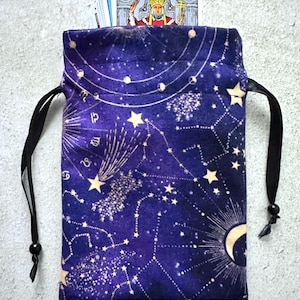 May include: A dark blue velvet tarot card bag with a celestial design featuring constellations, stars, and a crescent moon. The bag has a drawstring closure with black cords and holds a deck of tarot cards.