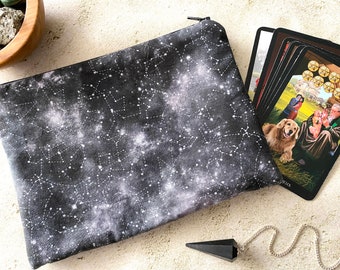 May include: A black fabric pouch with a zipper closure and a white constellation print. The pouch is lying on a light beige surface next to a deck of tarot cards and a black crystal pendulum.