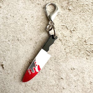 May include: A silver-colored charm with a lobster clasp, featuring a miniature knife pendant. The knife has a white blade with red blood-like splatters and a black handle. The charm is designed to be attached to a keychain or bag.