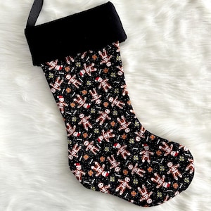 May include: A Christmas stocking featuring a black cuff and a black body patterned with gingerbread skeleton figures wearing red Santa hats, interspersed with small colorful peppermint candies and white bone shapes on a dark background.