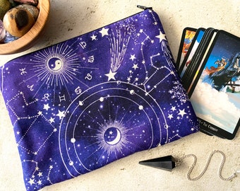 May include: A purple zippered pouch with a white celestial design featuring constellations, stars, and a crescent moon. The pouch is lying on a white surface with a black pendulum and a deck of tarot cards.