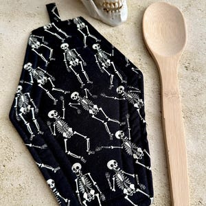 May include: Black coffin-shaped pot holder with a white skeleton pattern. A wooden spoon and a decorative skull are also in the image. The pot holder has a hanging loop. Halloween kitchen decor.