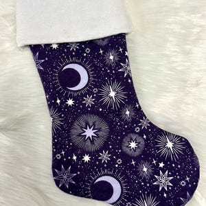May include: A Christmas stocking with a celestial design. The stocking features a dark purple background with a pattern of white stars, moons, and starbursts. The cuff is white, and a white ribbon loop is attached.