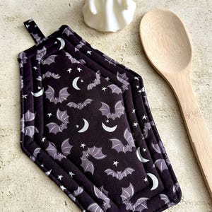May include: A black coffin-shaped pot holder with a repeating pattern of gray bats, white stars, and crescent moons. A small white ghost figurine and a wooden spoon are also in the image. The pot holder has a loop for hanging.