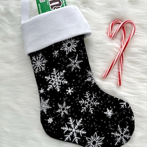 May include: A black Christmas stocking with white snowflake patterns and a white cuff. The stocking is filled with a green box of M&M's and is next to two red and white candy canes.