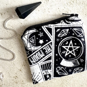 May include: Black and white fabric pouch with a zipper closure. The pouch features a repeating pattern of stars, moons, crystals, and a crystal ball with a pentagram. A black pendulum with a silver chain is also pictured.