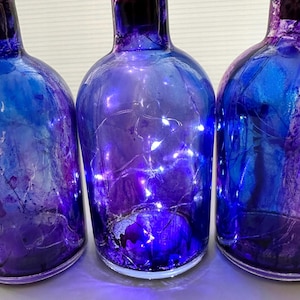 May include: Three decorative glass bottles with a gradient of blue and purple hues. Each bottle has a dark cork and is illuminated with small white lights inside. The bottles are arranged closely together, creating a visually appealing display.