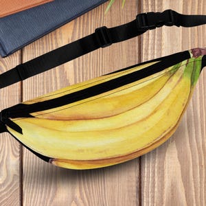 May include: A black fanny pack featuring a watercolor illustration of a bunch of ripe bananas. The pack has a black adjustable strap and buckle. The background is a wooden surface.