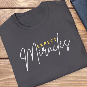 May include: A dark gray t-shirt with the text "Expect Miracles" in white cursive lettering with the word "Expect" in yellow.