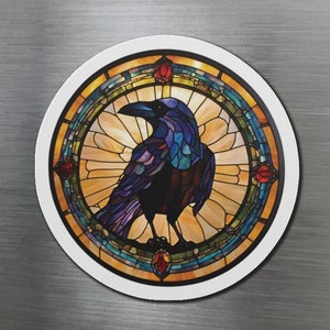 May include: A round magnet featuring a stained glass design of a raven with blue, purple, and black feathers. The raven is surrounded by a yellow, orange, and green stained glass design with red accents.