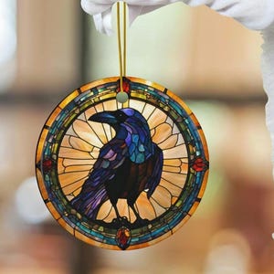 May include: A round, stained glass ornament featuring a black raven with blue and purple accents. The raven is perched in the center of a circular stained glass design with yellow, orange, and red accents.