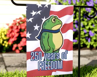 May include: A garden flag featuring a cartoon frog wearing a blue scarf, set against an American flag background. The flag reads "250 years of FREEDOM". The flag is displayed on a black metal stand, with green grass and colorful flowers in the background.