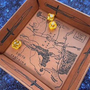 May include: A brown leather dice tray with a map design and black sword illustrations. The tray holds three yellow dice. The map includes text such as "South Farthing" and "The Shire".