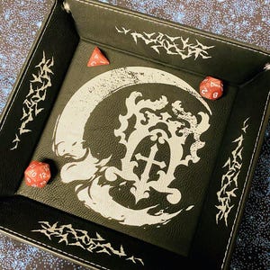 May include: Black square dice tray with a white gothic design and three red dice. The tray has a stitched border and decorative white accents. The dice are a triangular D4 and two D12s.