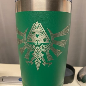 May include: A green insulated tumbler with a silver design featuring a triangle and stylized wings. The tumbler has a silver rim and a clear lid. The design is a popular video game symbol.