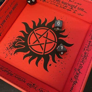 May include: A red dice tray with a black pentagram design and the text "Carry on my wayward son". Three dice are inside the tray. The tray has stitched edges and additional text around the border.
