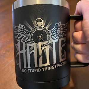 May include: Black travel mug with a silver rim and handle. The mug features a silver graphic of a compass with wings and the word "Haste" above the phrase "Do Stupid Things Faster."