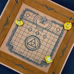May include: A square, brown leather dice tray with a grid and Zelda-themed artwork in the center. Three yellow dice are inside the tray. The tray's border features sword and Triforce designs. The tray is on a dark blue surface.