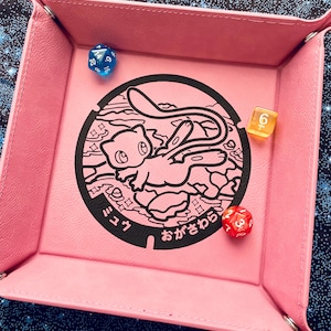 May include: A pink, square dice tray with a black graphic of a cartoon character and Japanese text. Three multi-sided dice in blue, yellow, and red are inside the tray. The tray has a stitched border.