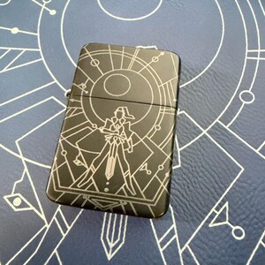 May include: A black metal lighter with a geometric design and a central figure holding a sword. The lighter has a silver-colored graphic and is set against a blue background with a white geometric pattern.