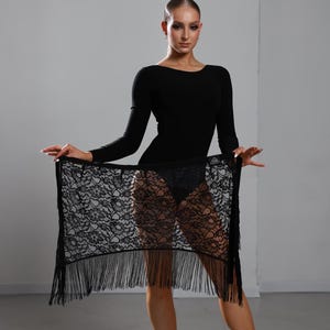 Puede incluir: Un body negro de manga larga combinado con una falda cruzada de encaje negro con un patrón floral y flecos largos en el borde inferior. El conjunto se completa con zapatos de baile de color beige con tiras finas y cierre de hebilla.