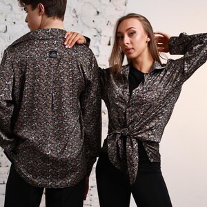 Può includere: Una camicia a maniche lunghe con bottoni caratterizzata da un motivo animalier ripetuto nei toni del marrone e del grigio. La camicia presenta un colletto, polsini abbottonati e una piccola etichetta rettangolare nera con testo bianco sul carré.