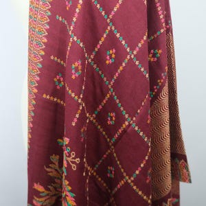 May include: Burgundy shawl with intricate geometric and floral patterns. The shawl features a diamond grid design with colorful accents in pink, yellow, green, and orange. The edges are adorned with detailed embroidery.