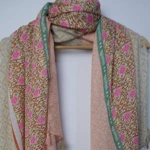 May include: A floral patterned scarf in shades of pink, brown, and beige hangs on a wooden hanger. The scarf features a repeating pink flower design on a brown background, with a light green border and tassel details.