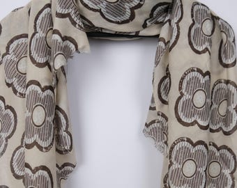 May include: A beige scarf with a repeating floral pattern in brown. The flowers have a circular center and striped petals. The scarf is draped over a black hanger, with the ends hanging down. The edges of the scarf are frayed.