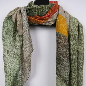 Op de afbeelding: Een sjaal met patroon in groen, bruin, geel en oranje hangt aan een zwarte hanger. De sjaal heeft een textuurontwerp met diagonale lijnen en kleurvlakken. De sjaal is gemaakt van een lichtgewicht materiaal.