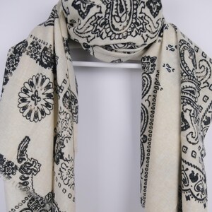 Könnte beinhalten: Cremefarbener Schal mit schwarzem Paisley-Muster. Der Schal ist auf einem weißen Kleiderbügel drapiert, der das detaillierte Muster zeigt. Die Kanten sind mit Fransen versehen, was dem Accessoire Textur verleiht.