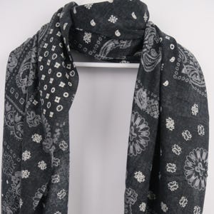 Puede incluir: Bufanda gris oscuro con un diseño de cachemir y floral blanco. Colgada de una percha blanca, muestra su forma rectangular y bordes deshilachados. La tela parece suave y ligera, adecuada para varios estilos.
