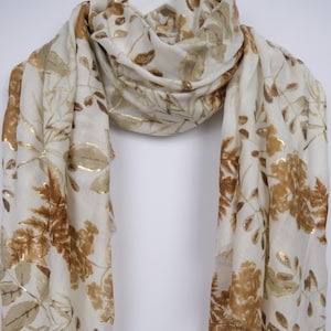 May include: A cream-colored scarf with a botanical pattern. The scarf features a repeating design of brown and gold leaves and foliage. The scarf is made of a lightweight fabric and is draped against a white background.