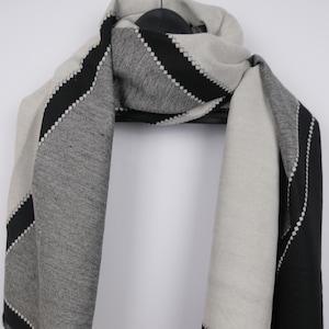 May include: A gray, black, and white scarf with a geometric design. The scarf has a fringed edge and a black border with white stitching. The scarf is draped over a black hanger against a white background.