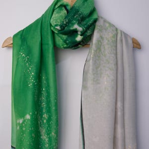 Op de afbeelding: Een groene en witte sjaal met een gouden glitterontwerp. De sjaal is gevouwen en hangt aan een houten hanger.