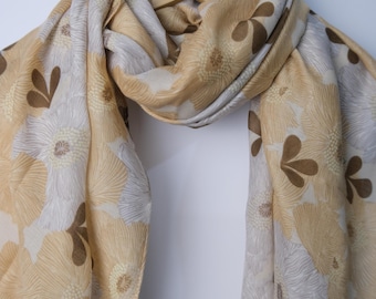 May include: A beige and gray floral scarf with a repeating pattern of stylized flowers and leaf shapes. The scarf is draped over a wooden hanger, showcasing its flowing fabric and design.