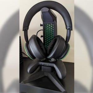 Könnte beinhalten: Ein schwarzes Gaming-Headset mit grünen Akzenten liegt auf einem schwarzen Controller-Ständer. Das Headset befindet sich auf einem Ständer, der einem Videospiel-Controller ähnelt.