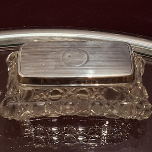 May include: A rectangular silver box with a textured lid and a decorative crystal base. The lid has a circular indentation in the center. The box is displayed on a reflective surface.
