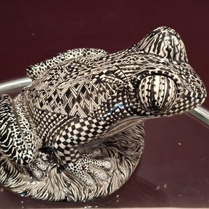 Pode incluir: Estatueta de rã de cerâmica com um padrão geométrico em preto e branco. A rã tem olhos grandes e detalhados e está pousada sobre uma base com um design texturizado. A estética geral é artística e decorativa.