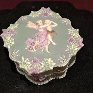 Pode incluir: Caixa de bugigangas decorativa de cerâmica com tampa verde clara com um design esculpido de duas figuras dançando. A caixa é adornada com detalhes florais roxos e brancos e uma borda ondulada.